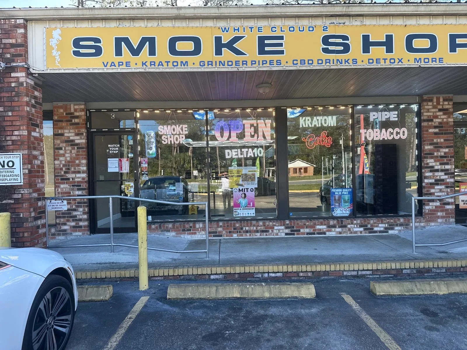 The yellow White Cloud 2 Smoke Shop sign by day, listing vape, kratom, grinder pipes, CBD drinks and detox